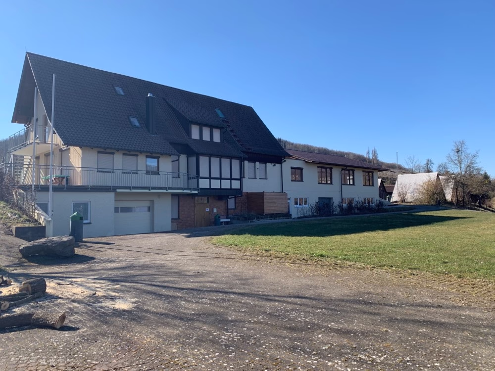 Mehrteiliger Betriebshof mit modernisiertem Wohn- und Gewerbegebäude, Satteldächern, Garagen und gepflastertem Vorplatz vor Grünfläche.