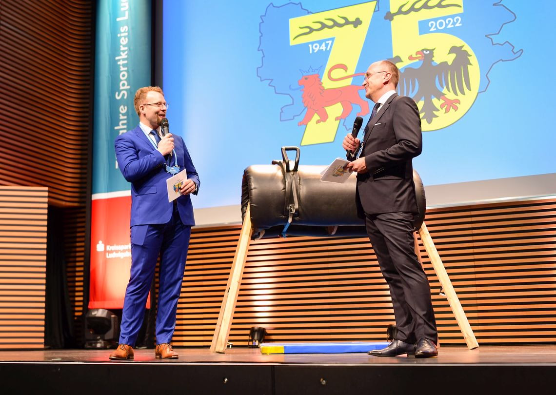 Zwei Redner stehen auf einer Bühne, halten Mikrofone und sprechen; im Hintergrund Banner und Sport-Motiv, im Vordergrund ein Podest mit Fußpodium.
