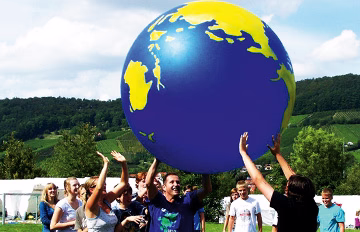 Kindergruppe im Freien wirft bzw. hebt einen großen, blauen Weltballon gemeinsam, symbolisiert gemeinsame Freizeit und Lernen durch Bewegung.