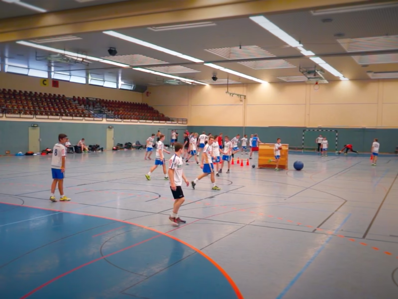 Mannschaftsszene in einer Sporthalle: mehrere Spieler und ein Ball bei einem Spiel, im Hintergrund Zuschauertribüne.