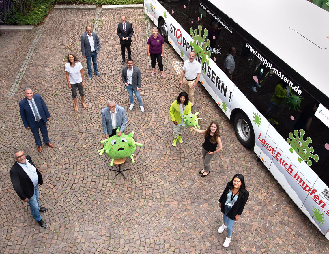 Gruppe von Menschen vor einem Werbemobil mit Stoppt Masern-Logo und grünen Maskottchen; Kampagnenmotiv auf der Fahrzeugseite.