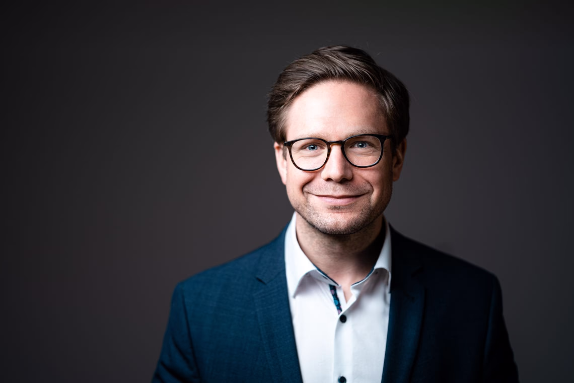 Porträt eines Mannes mit Brille im Anzug, frontal ausgerichtet vor einfarbigem Hintergrund, freundlicher Gesichtsausdruck.