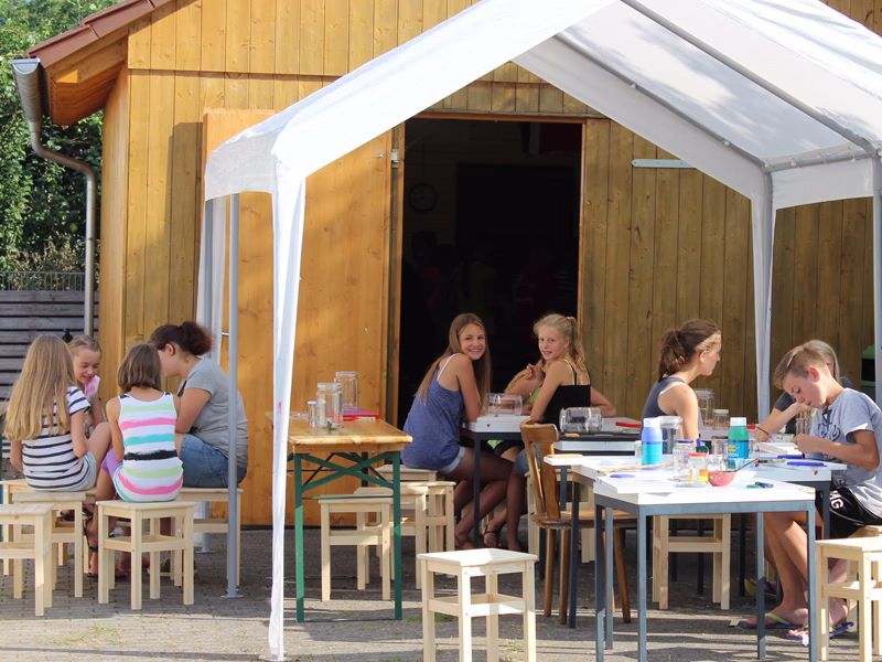 Kinder und Erwachsene sitzen an Tischen in einer hölzernen Bastelhütte unter einer transparenten Zeltplane und basteln gemeinsam.
