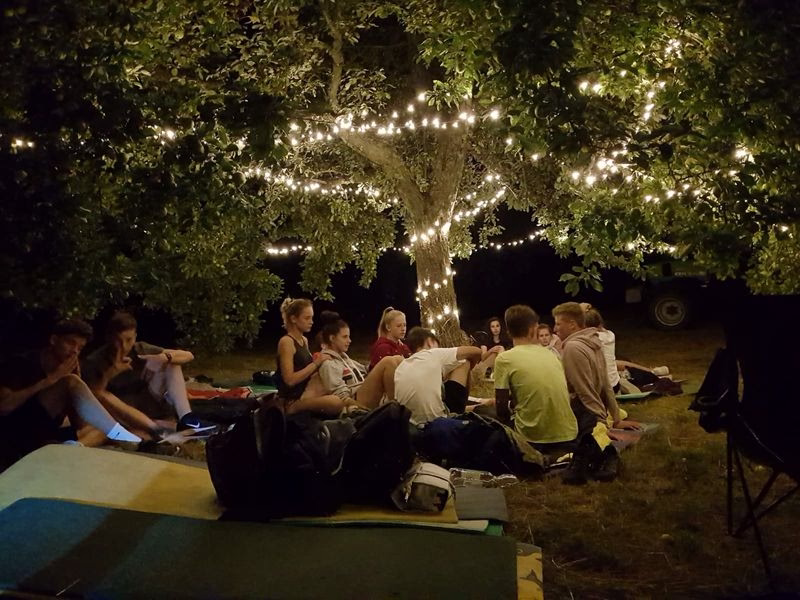Gruppe sitzt im Garten um eine Tischfläche, unter Bäumen mit Lichterkette—outdoor Veranstaltungsszene bei Abendlicht.