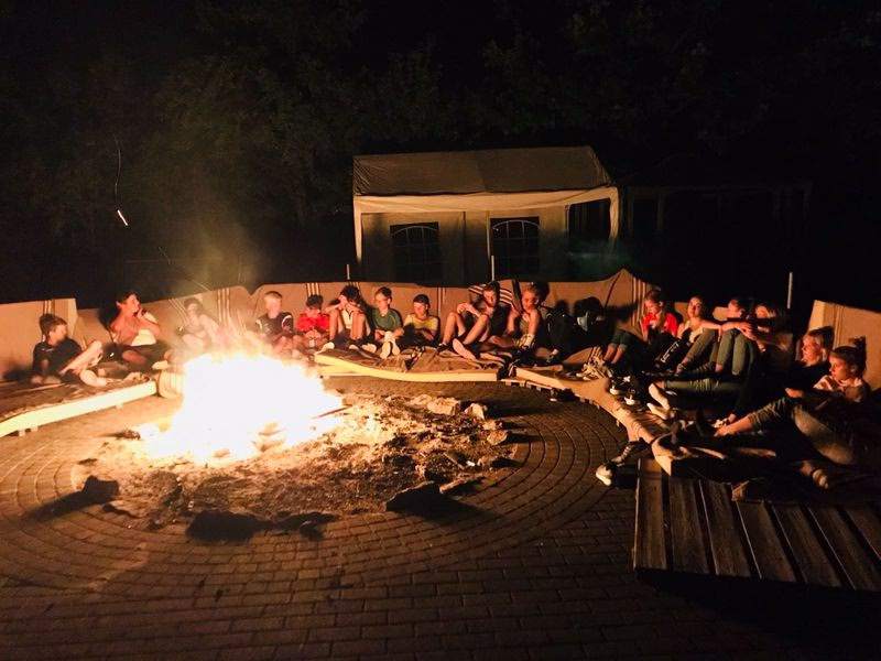 Gruppe sitzt am Lagerfeuer vor einer beleuchteten Festzelthütte, einige schauen ins Feuer, andere essen/essen gemeinsam.