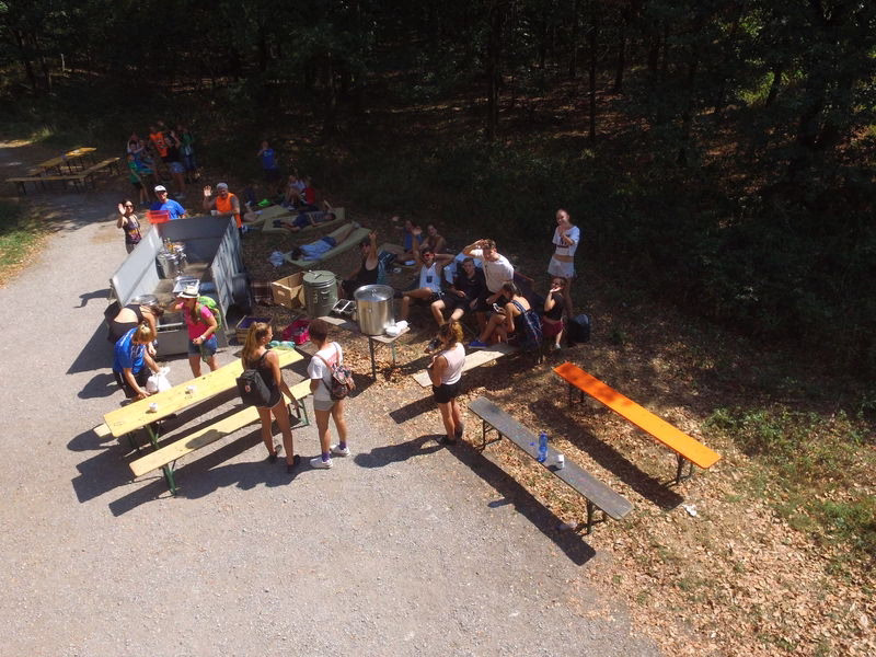 Grill- und Essenstafel im Freien: Menschen stehen und sitzen rund um ein großes Grillgerät auf dem Wanderparkplatz.