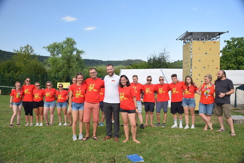 Gruppe von Teilnehmenden im roten Teamshirt steht auf Rasen und lächelt vor einer Kletterwand und einer gespannten Plane.