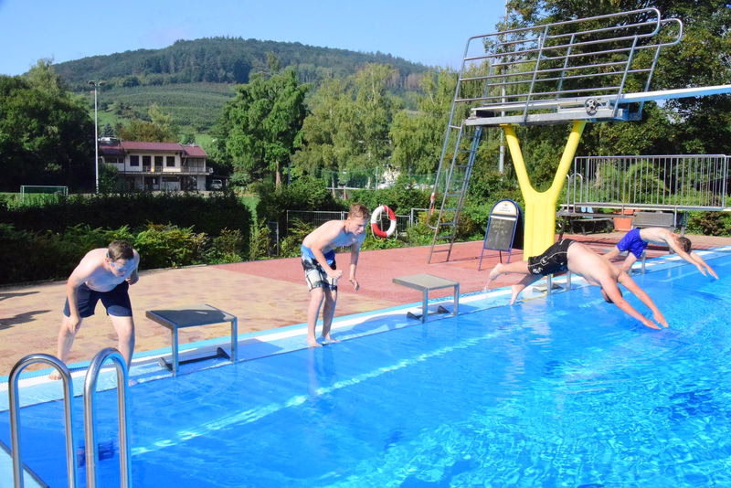Außenpool mit vier Springerinnen und einem Wasser-Entertainment-Gerät; im Hintergrund Bäume und Hügel, genutzt zum Schwimmen und Spaß im Freien.