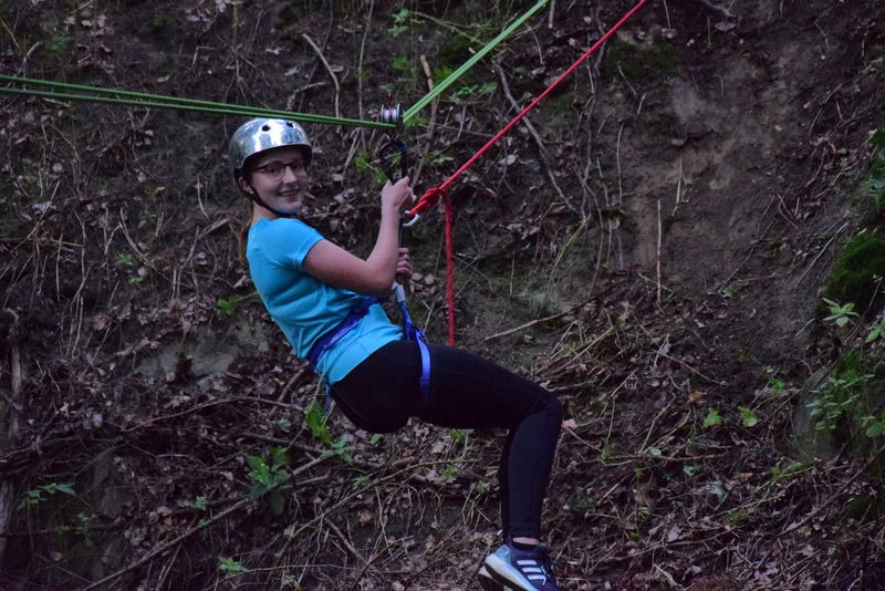 Kind mit Kletterhelm im Waldseilgarten, kniend und mit Klettergriff an den roten Seilen gesichert.