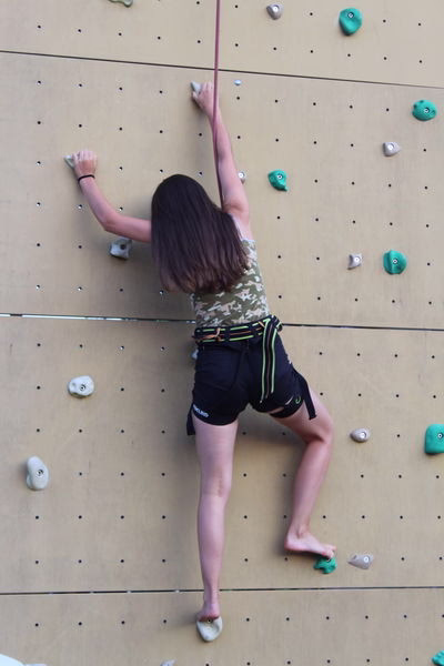 Kletterin an einer mit Griffen versehenen Boulderwand; sie streckt den Arm nach oben, um den nächsten Tritt zu greifen.