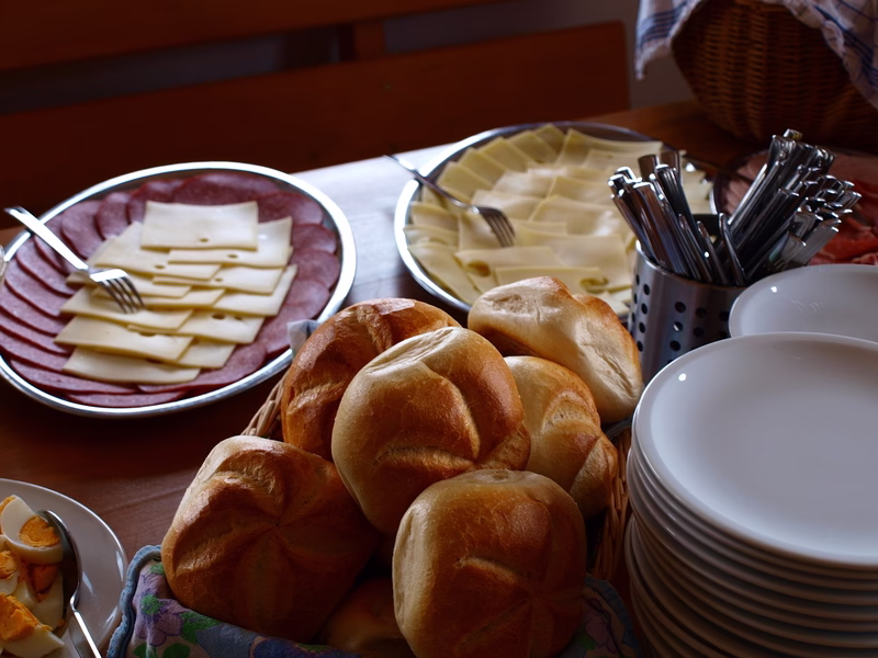 Teller mit Käseplatten, Brötchen und Servietten, daneben Besteck im Becher; gedeckter Frühstückstisch mit verschiedenen Snacks.