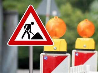 Warnschild mit Fußgängerbereich und Dreiecksymbol; im Hintergrund Baustellenabsperrungen und orangefarbene Scheinwerfer/Marker.