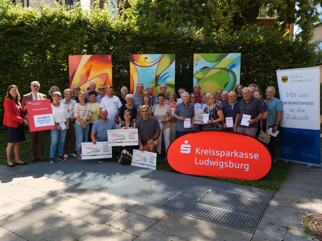 Gruppenbild vor bunten Wandbildern im Garten: mehrere Personen posieren mit Urkunden und Schecken, vor einer großen roten „Kreissparkasse“-Bankmarke.