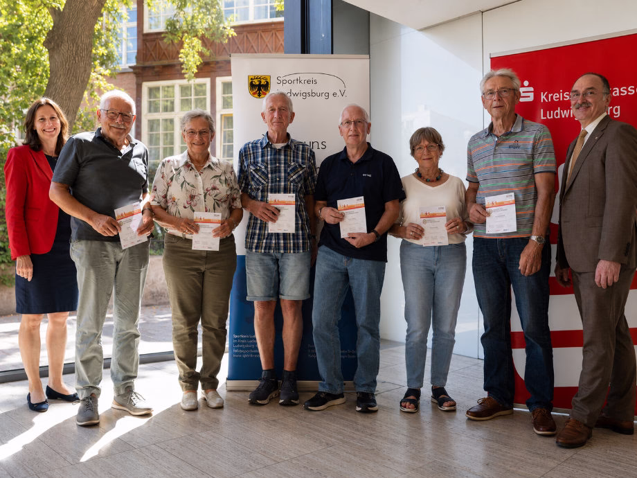 Gruppe älterer Personen hält Urkunden bei einer Veranstaltung; sie stehen nebeneinander im Eingangsbereich als Zeichen von Ehrungen.