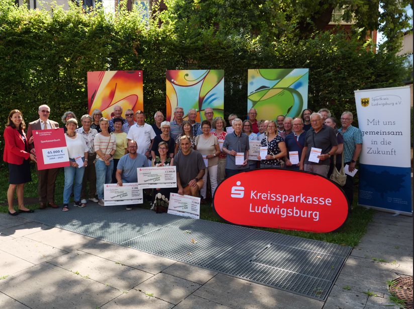 Gruppenfoto im Freien: mehrere Personen halten Schecks und Urkunden vor einem großen Logo-Schild; dient als Anlassfoto für einen Spenden-Event.
